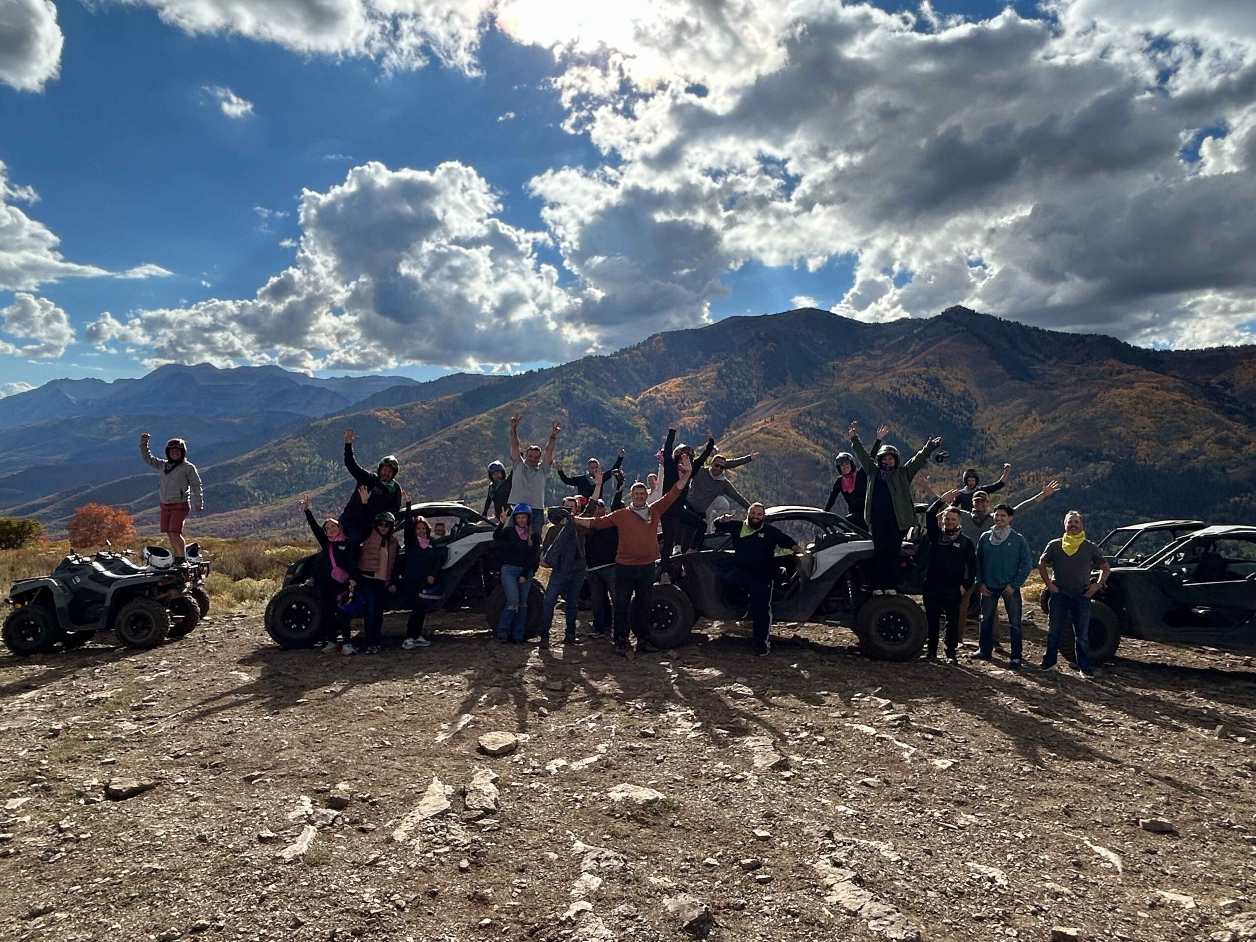 Group of people with helmets standing and raising hands beside parked atvs in a mountainous landscape under a cloudy sky