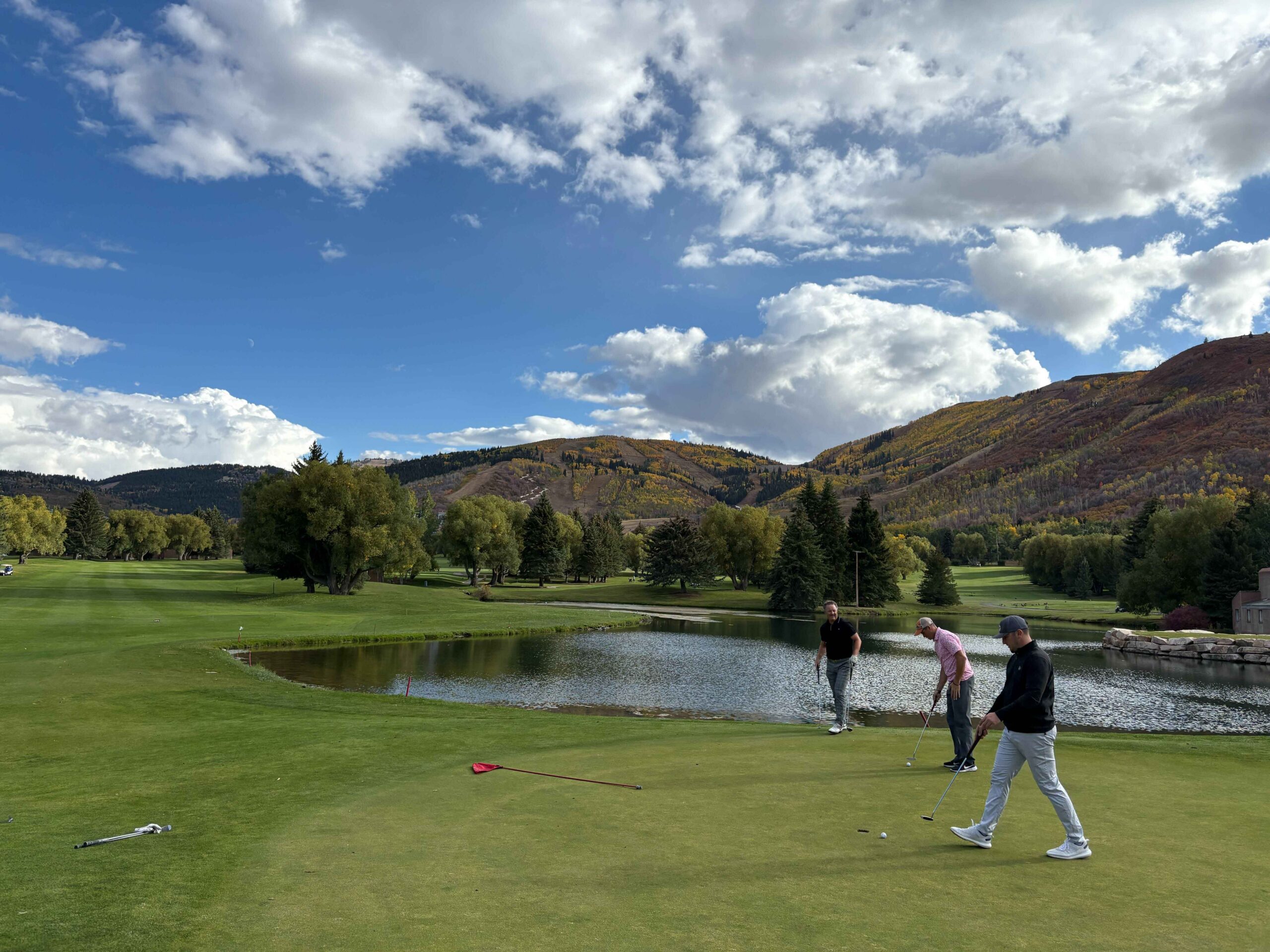Golfers walking on a green golf course beside a small pond with autumn trees and a hillslope in the background under a blue partly cloudy sky
