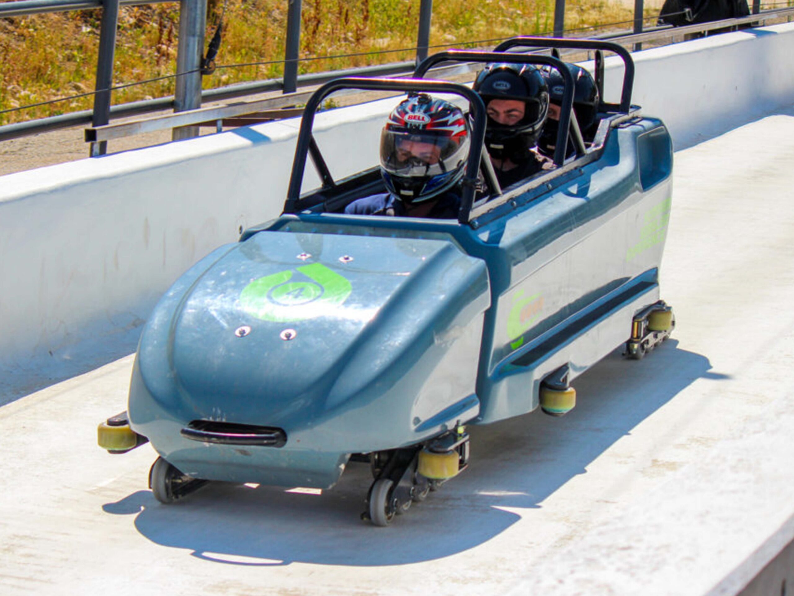 Two riders wearing helmets sit in a blue amusement ride car on a track with a railing beside them