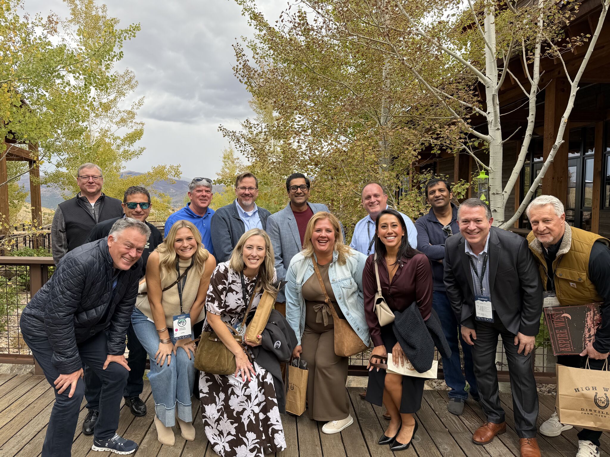 Group of adults posing for a photo on a wooden deck with trees and a building in the background smiling at the camera and wearing badges